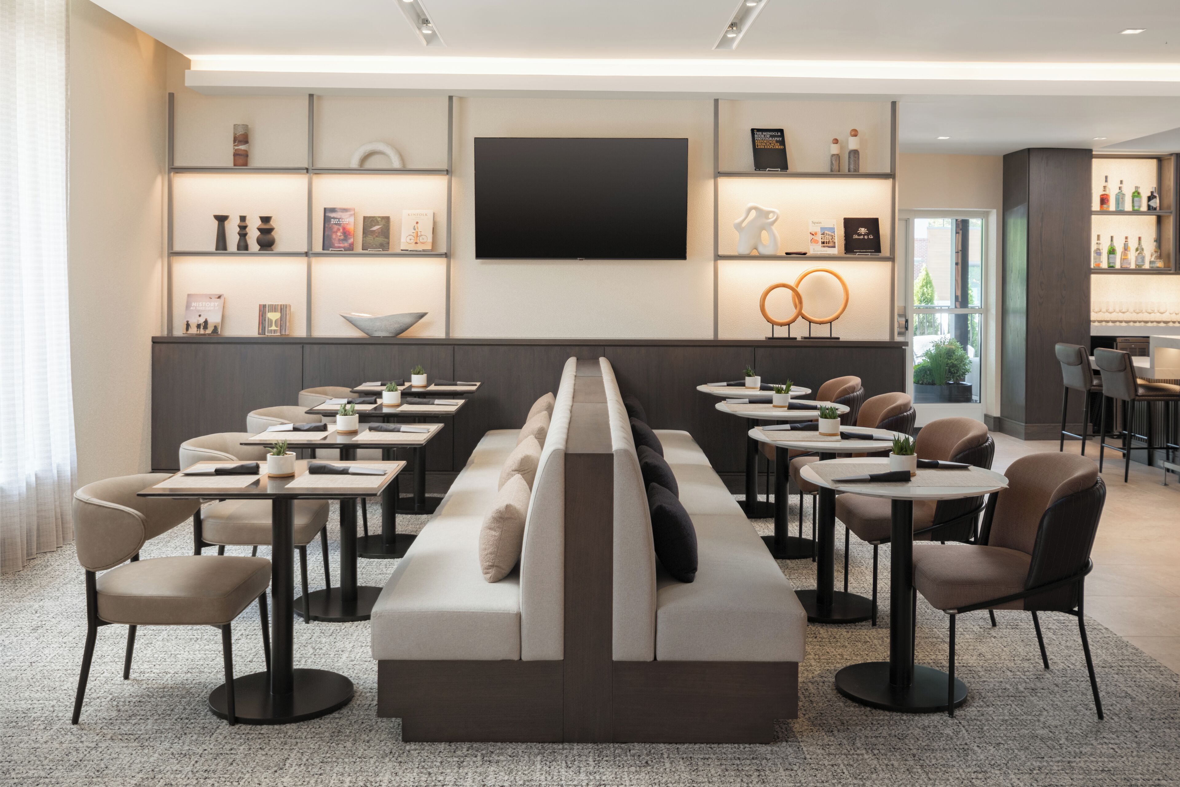 Un espace de détente moderne comprenant un long banc, plusieurs tables et chaises, ainsi qu’un grand téléviseur à écran plat et des étagères remplies de livres et d’objets décoratifs.