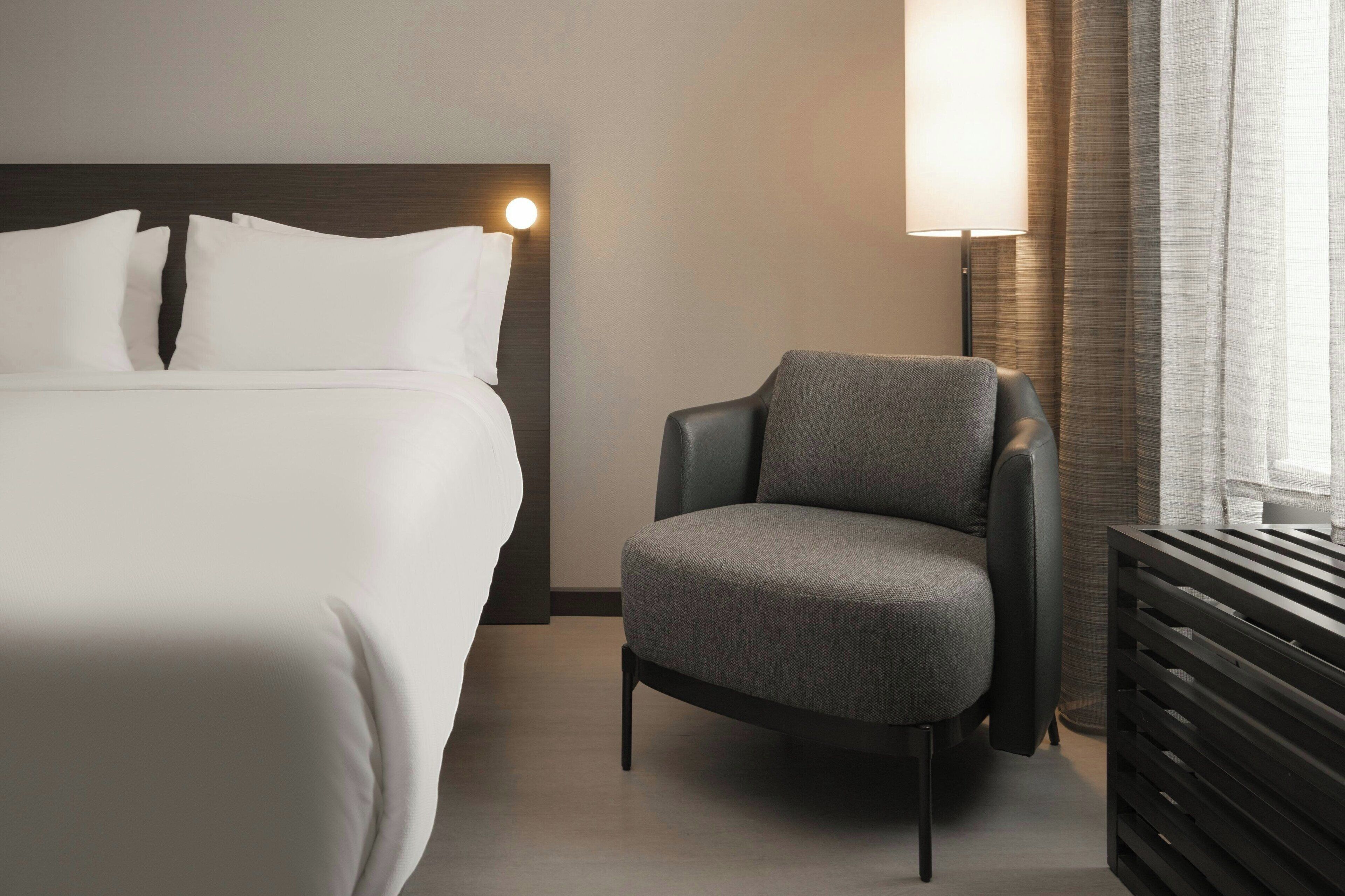 A hotel room with a large bed, a grey armchair, a bedside table, and a lamp.