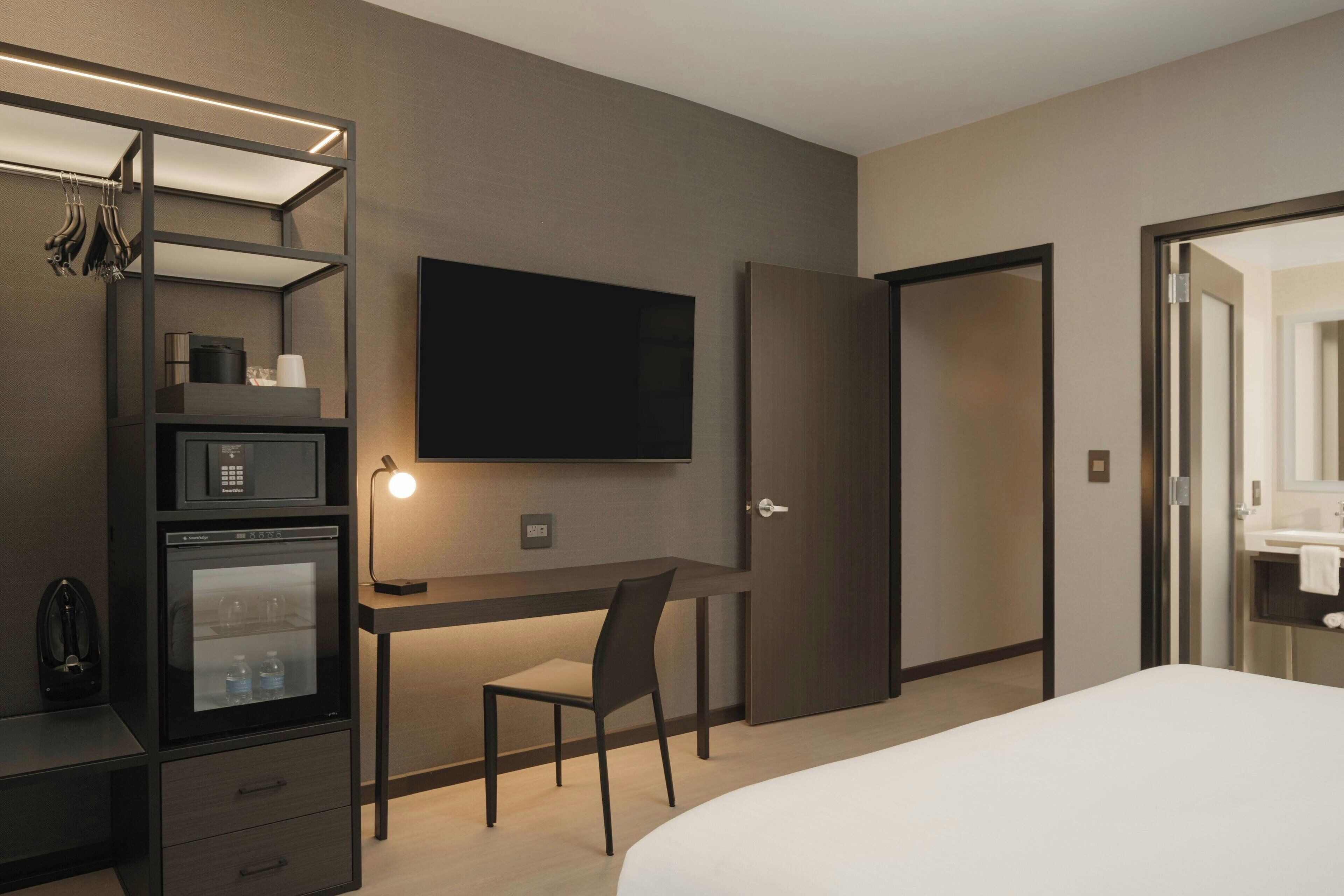 A hotel room with a bed, a desk with a chair, a television, a minibar, and a door leading to the bathroom.