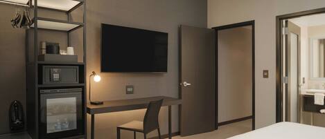 A hotel room with a bed, a desk with a chair, a television, a minibar, and a door leading to the bathroom.