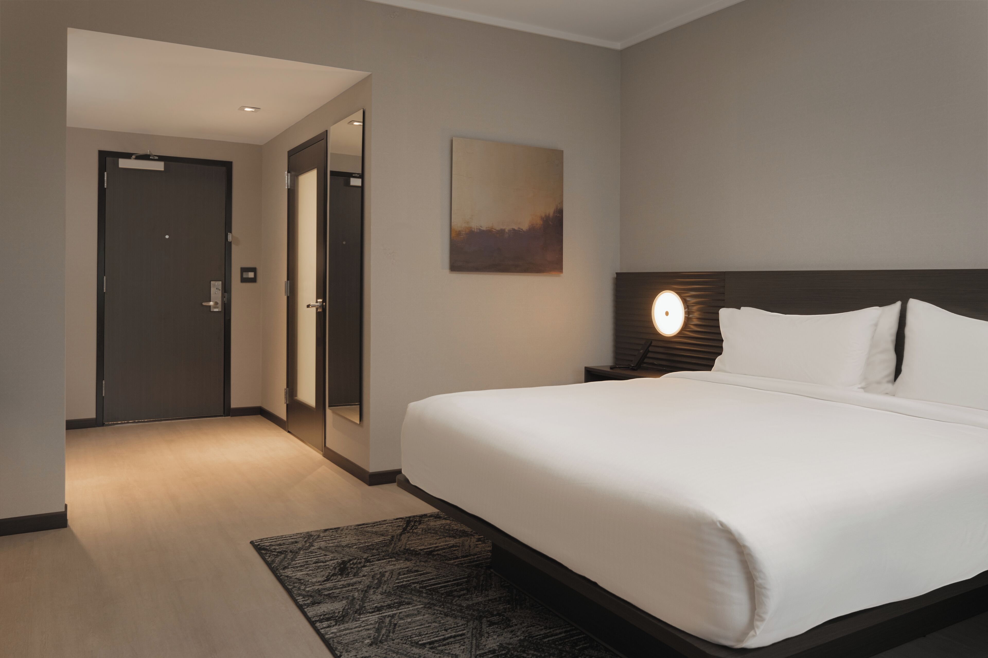 Hotellihuone, jossa on suuri sänky, yöpöytä lampun kanssa, taulu seinällä ja ovi, joka johtaa toiseen huoneeseen.