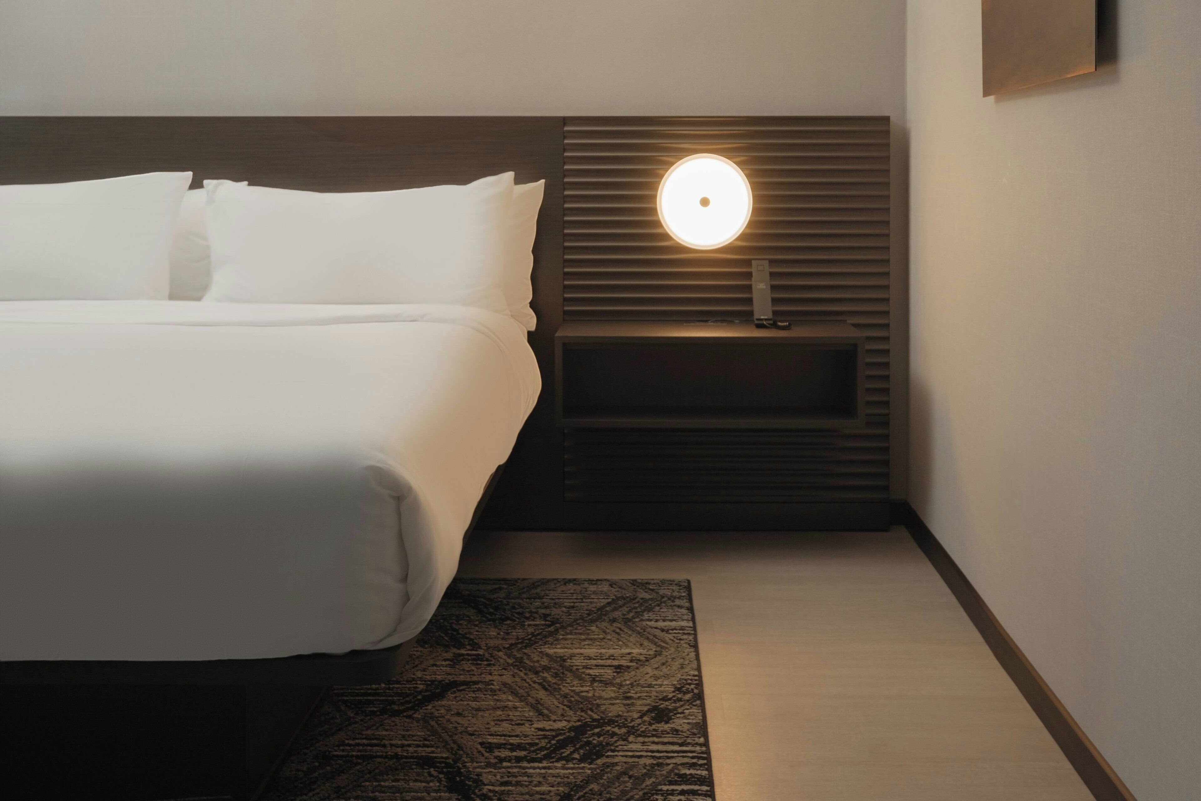 Una cama bien tendida con sábanas blancas, una mesita de noche con una lámpara moderna y una alfombra estampada sobre un piso de madera.