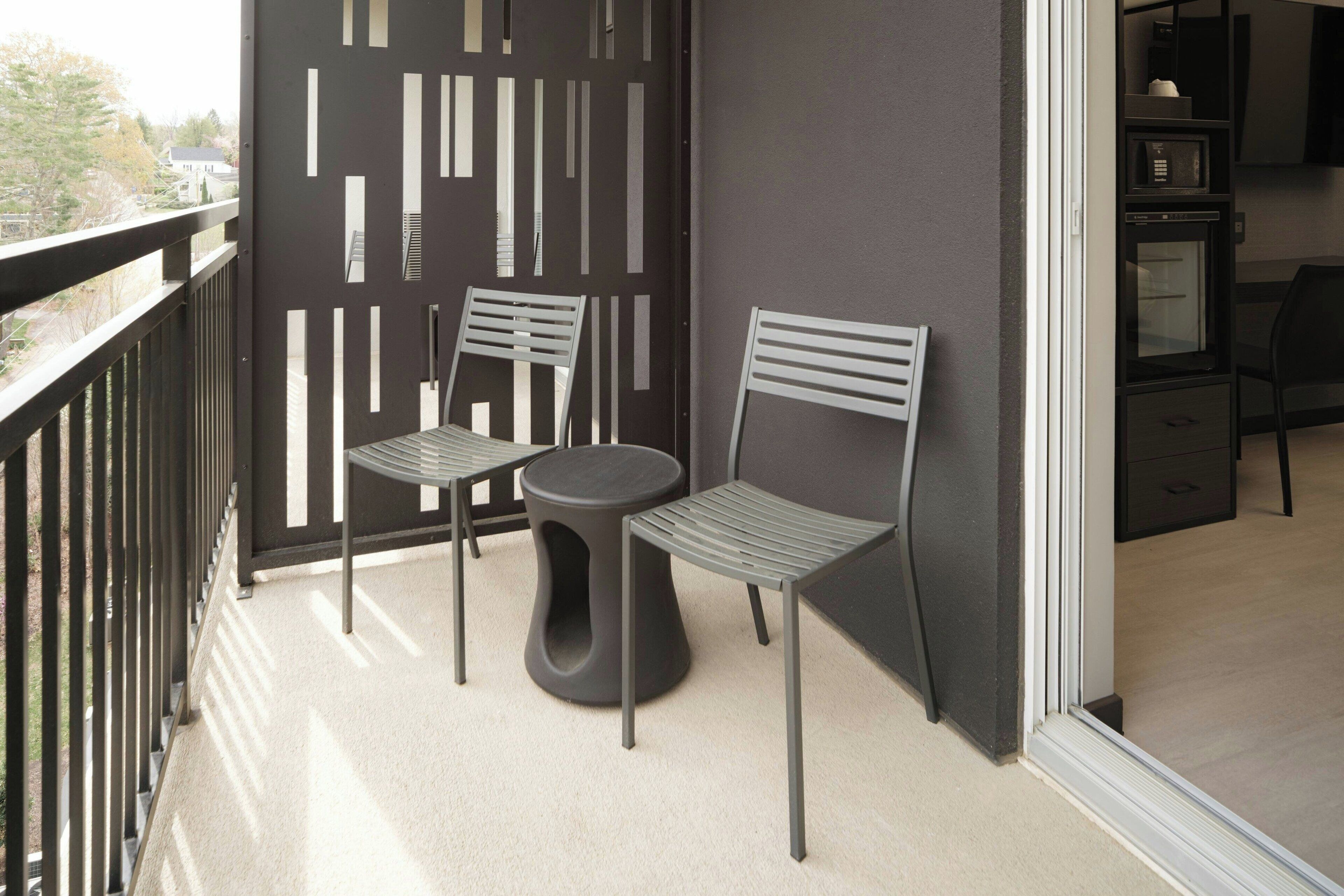 Un balcon avec deux chaises et une petite table, un mur sombre orné d’un motif géométrique et une vue sur l’extérieur.