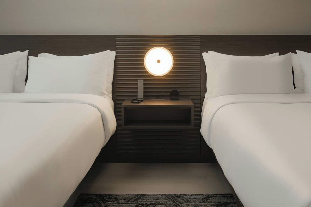 A hotel room with two beds, a bedside table with a lamp, and a wall-mounted light fixture.