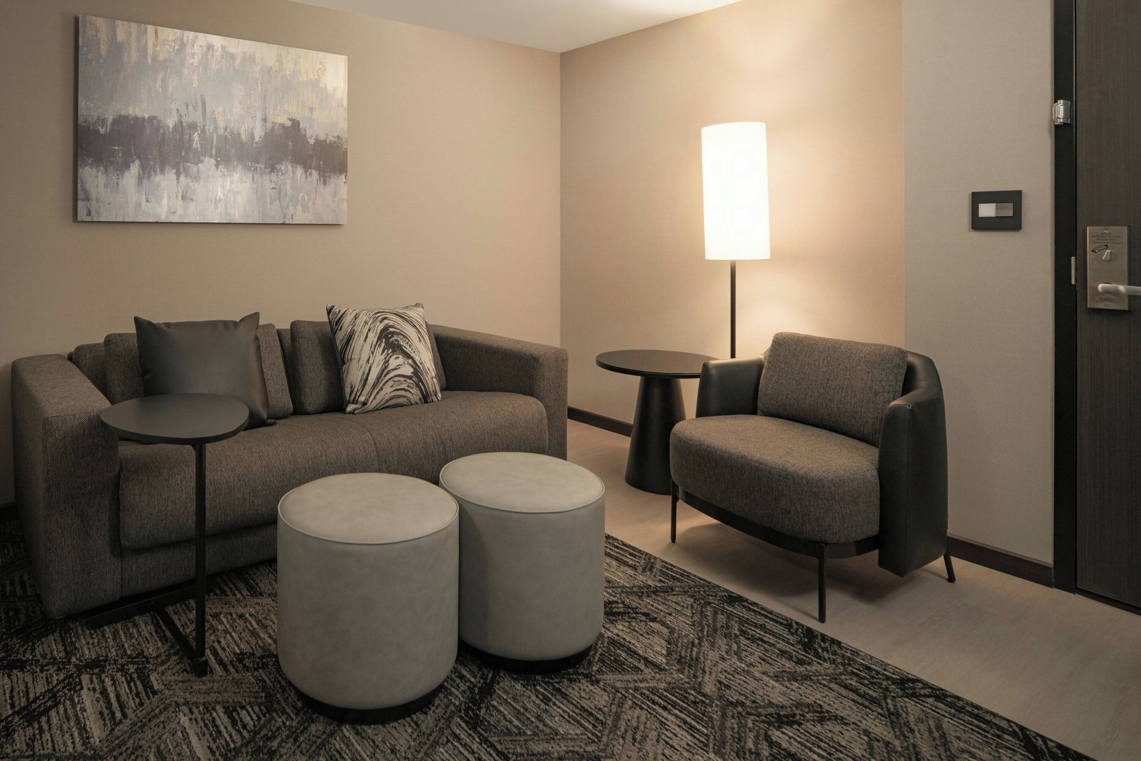 Ein modernes Hotelzimmer mit einem grauen Sofa, zwei Hocker, einem runden Tisch und einer Lampe.
