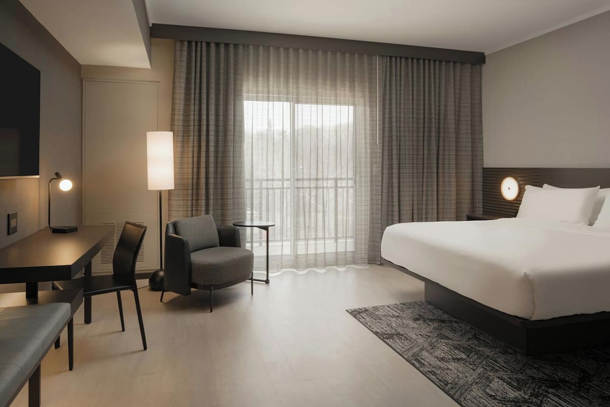 A hotel room with a bed, a desk, a chair, a lamp, and a view of the outdoors through the window.