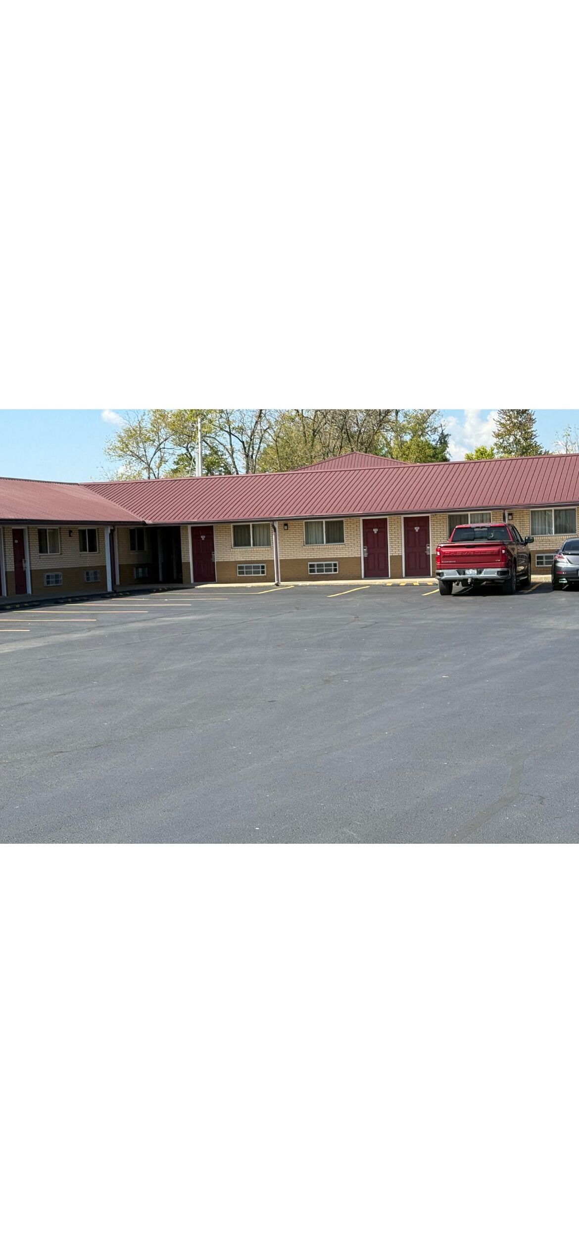 A motel with a red roof and beige walls, featuring a parking lot and a red pickup truck parked in front.