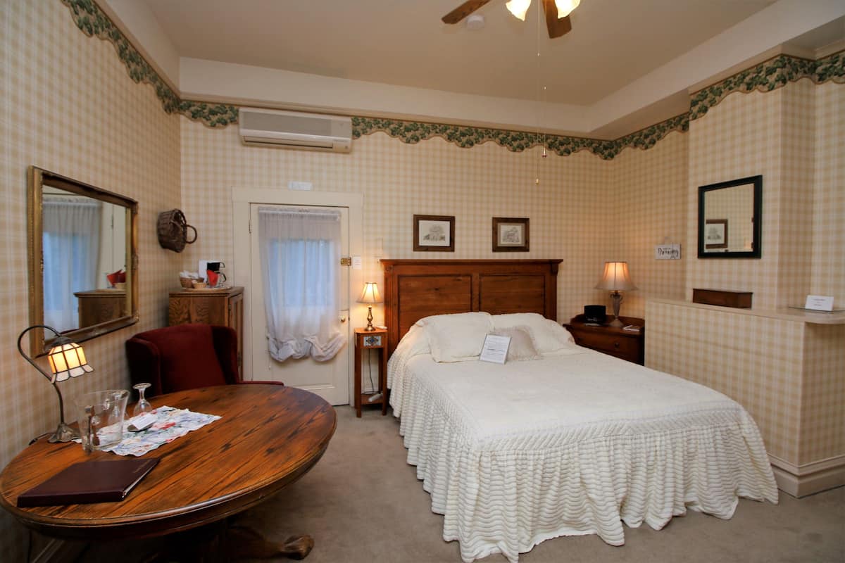 A hotel room with a large bed, a wooden desk, a chair, a small table, a mirror, and a ceiling fan.