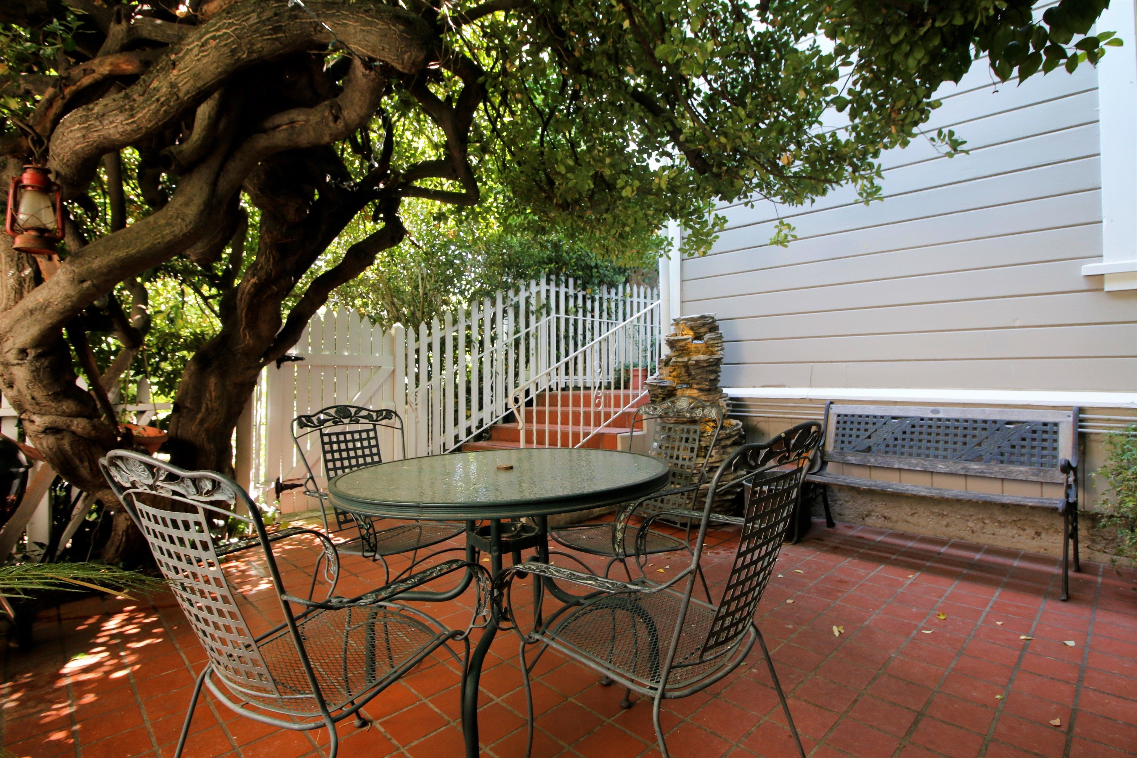 An outdoor patio with a round table and metal chairs, surrounded by a white picket fence and a tree.