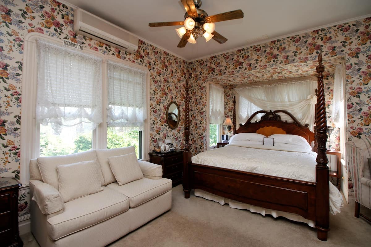 A bedroom with a four-poster bed, a white sofa, floral wallpaper, and a ceiling fan.