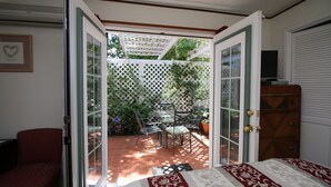 A bedroom with an open door leading to a patio with a table and chairs, a wooden dresser, and a red chair. - Bradford Place Inn & Gardens (Sonora)