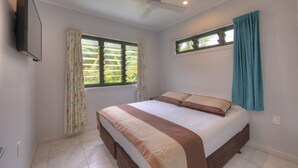 Un dormitorio con cama, dos ventanas con cortinas, ventilador de techo y televisor montado en la pared.