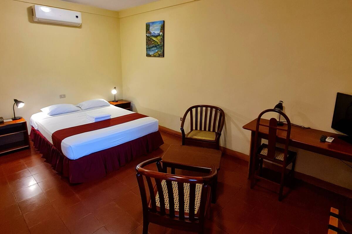 A hotel room with a bed, two chairs, a desk, a TV, and an air conditioning unit on the wall.