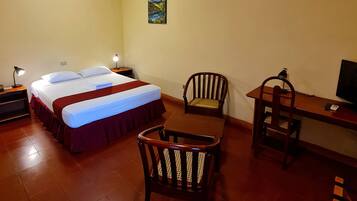 A hotel room with a bed, two chairs, a desk, a TV, and an air conditioning unit on the wall.