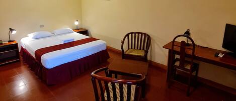 A hotel room with a bed, two chairs, a desk, a TV, and an air conditioning unit on the wall.