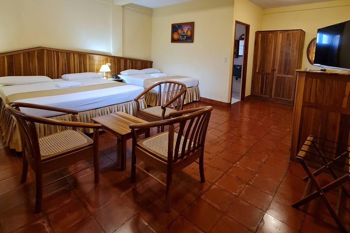 A hotel room with two beds, a wooden dining table, chairs, a television, and a cabinet.