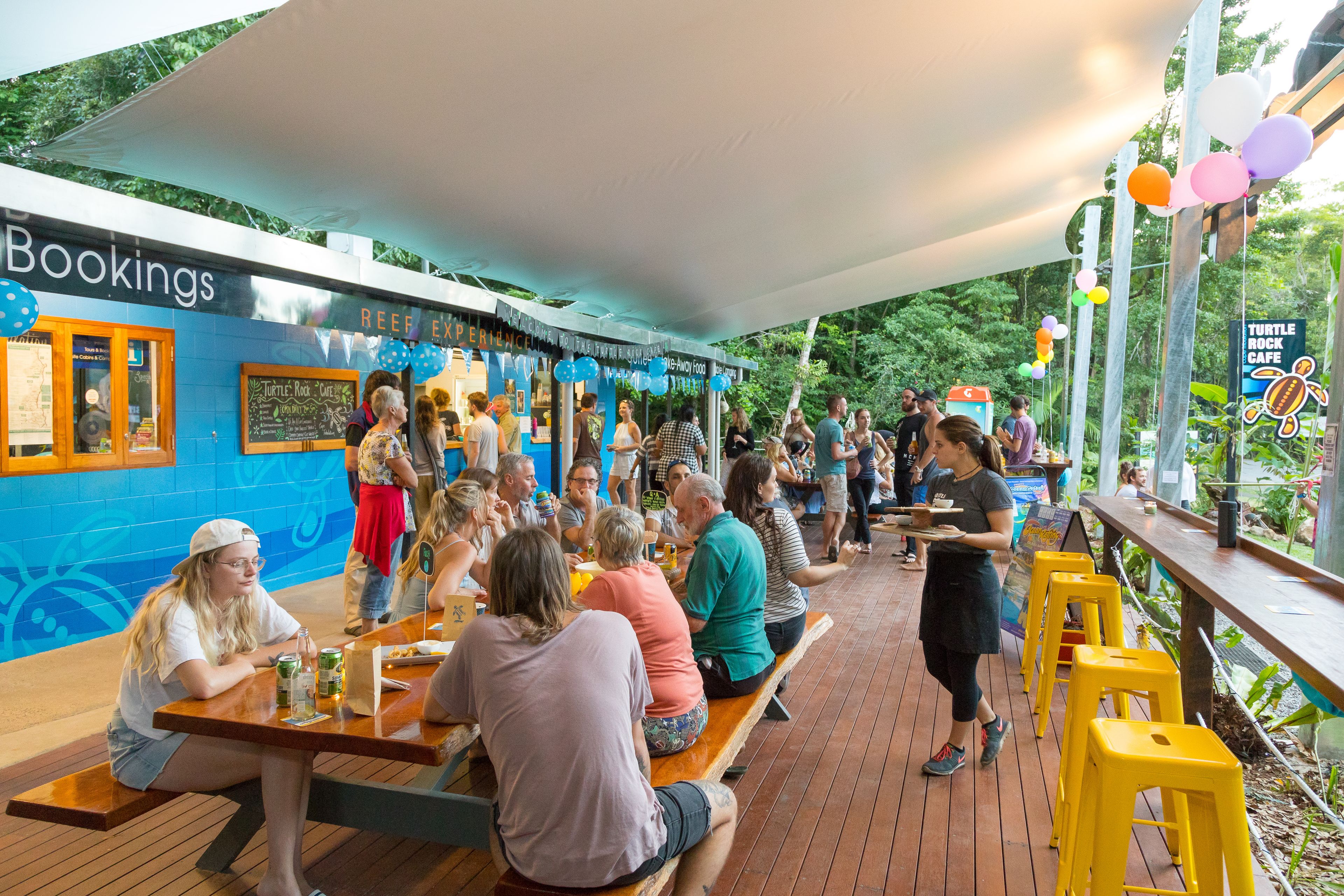 An outdoor seating area with wooden tables and benches, a blue wall with a 'Bookings' sign, and a 'Turtle Rock Cafe' sign.