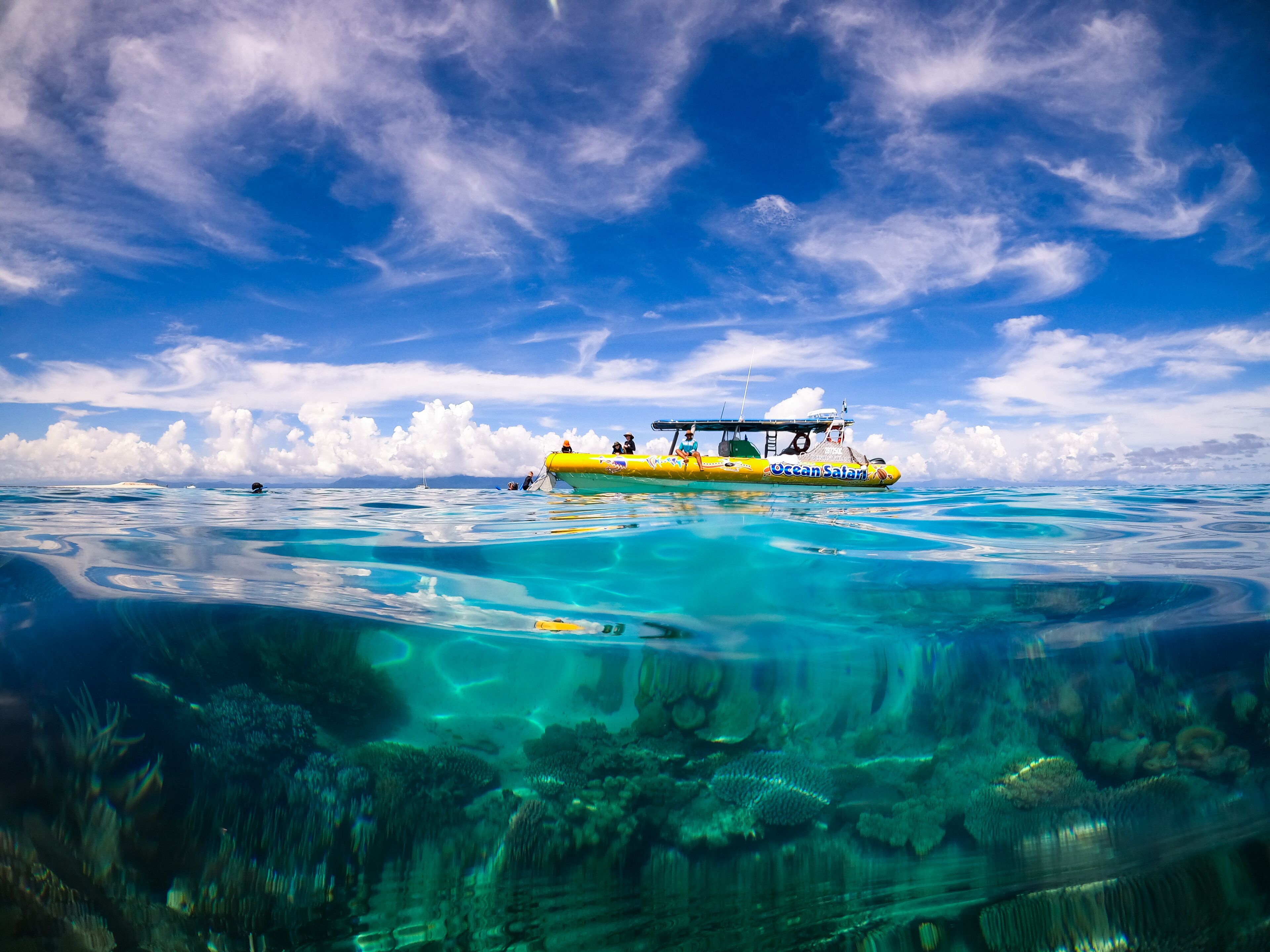A yellow boat with "Ocean Safari" written on it, floating on clear blue water with a view of the sky and clouds above.