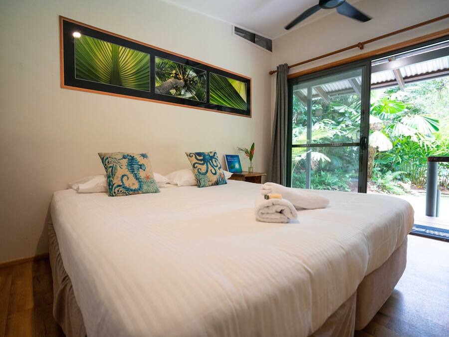 A well-lit bedroom with a large bed, a ceiling fan, and a view of lush greenery through a sliding glass door.
