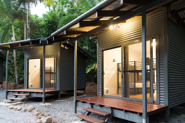 Duas cabines modernas, com paredes de vidro e decks de madeira, além de grades metálicas, localizadas em ambiente tropical com vegetação visível.