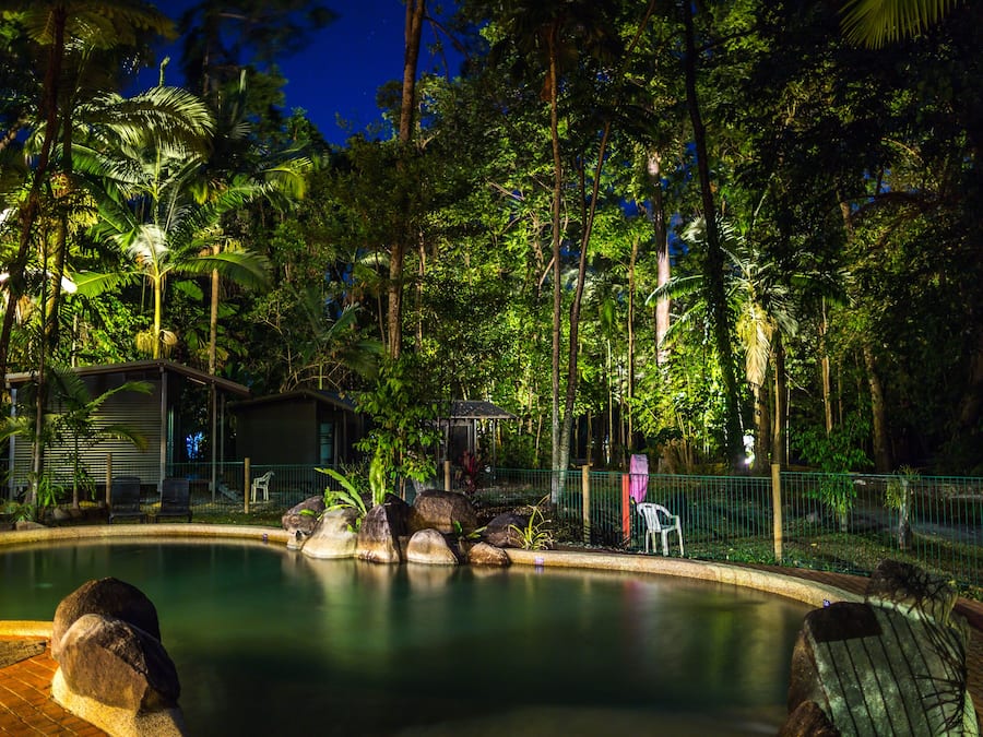 A swimming pool surrounded by lush greenery and trees, with a small building and a fence in the background.