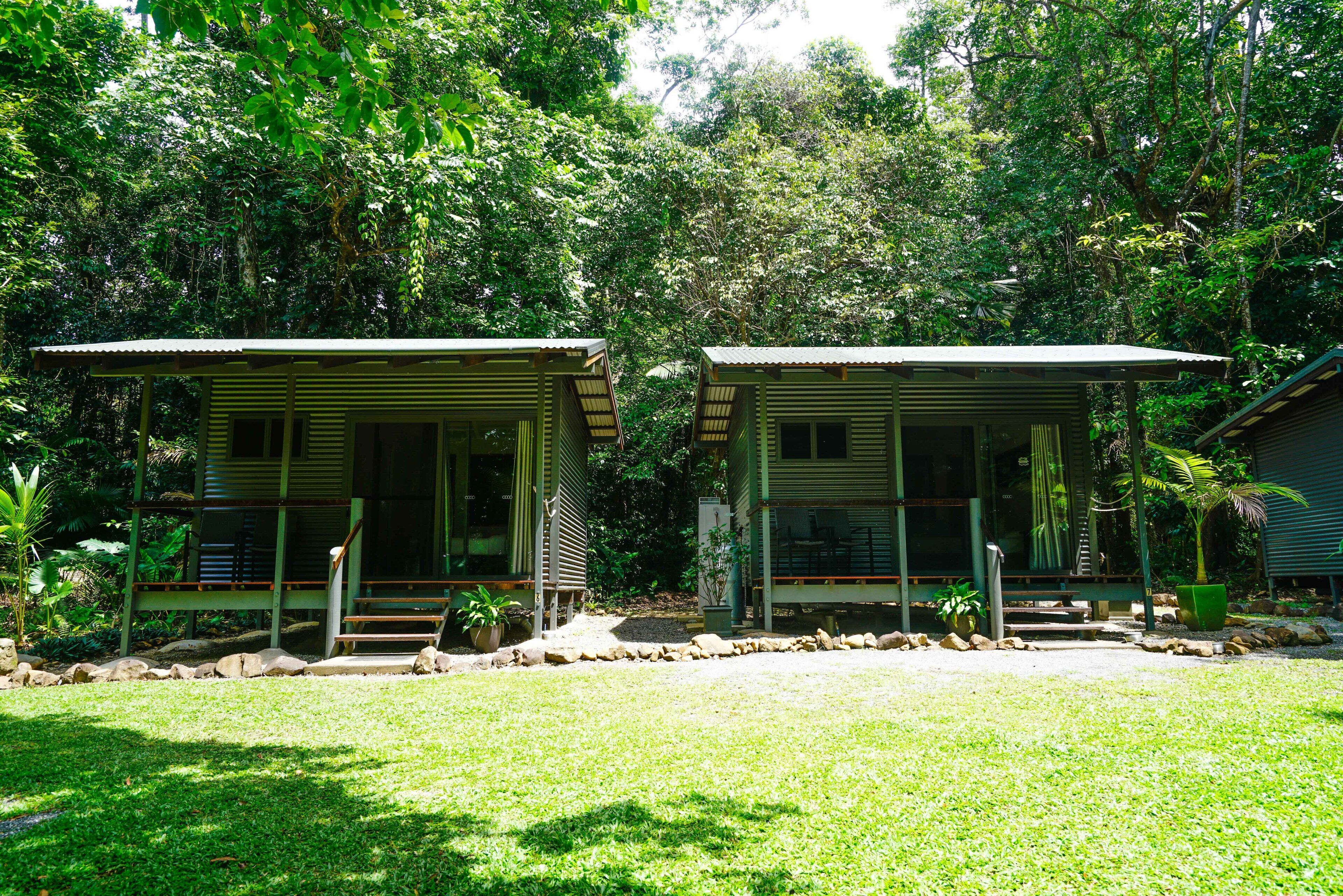 Two small, single-story cabins with sloped roofs, surrounded by lush greenery and a well-maintained lawn.