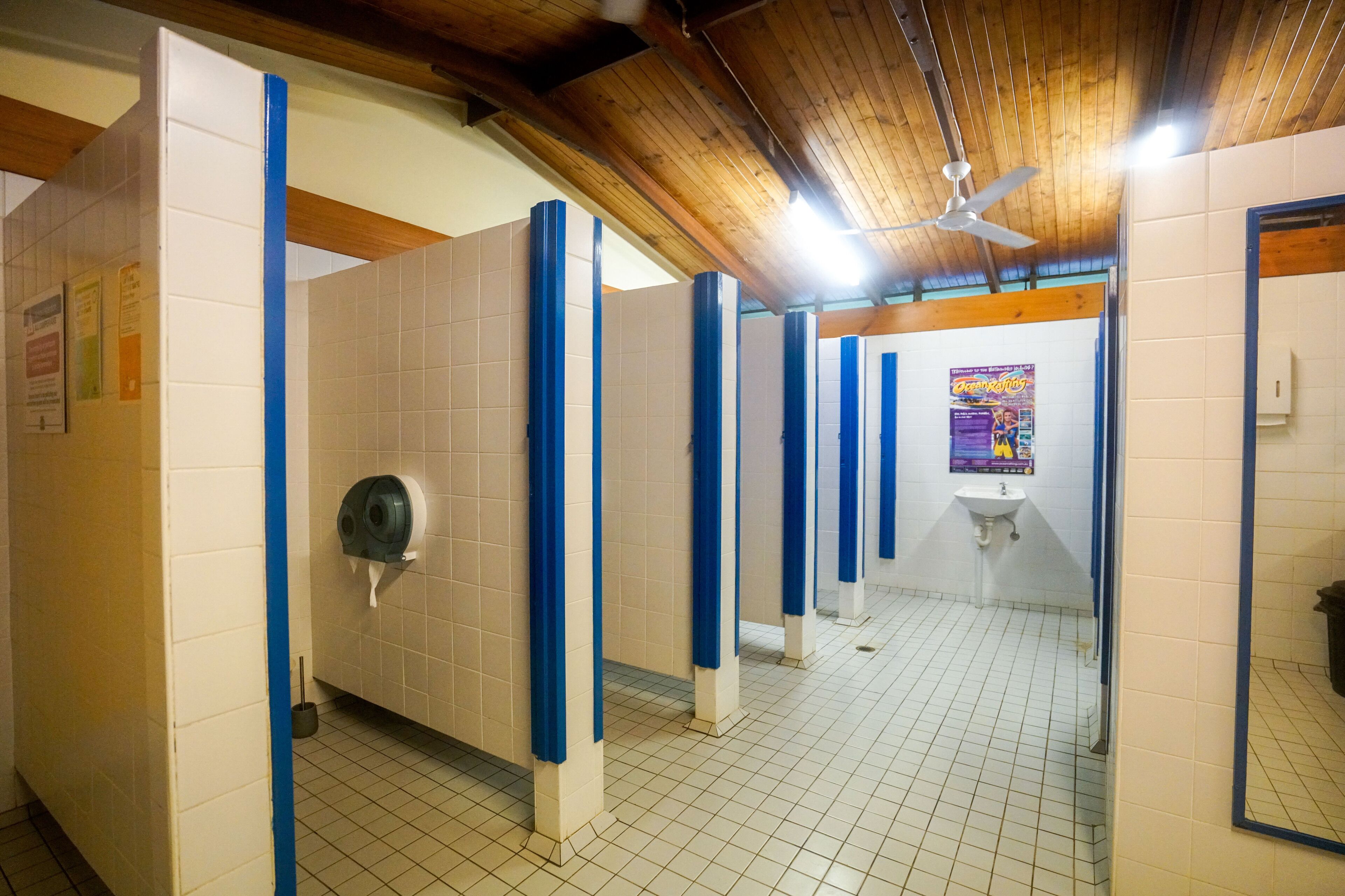 A public restroom with blue partitions, a hand dryer, a sink, and a ceiling fan.