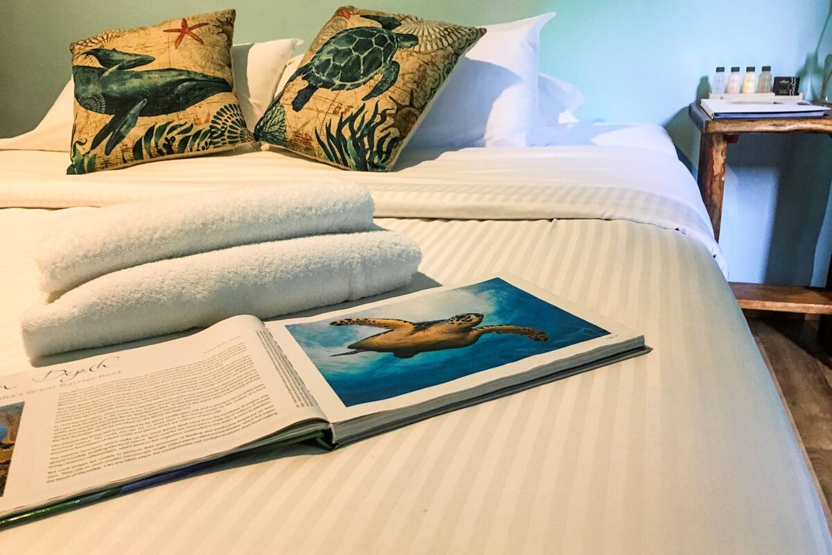Ein ordentlich bezogenes Bett mit Kissen, einem Buch und einem kleinen Tisch mit Flaschen.