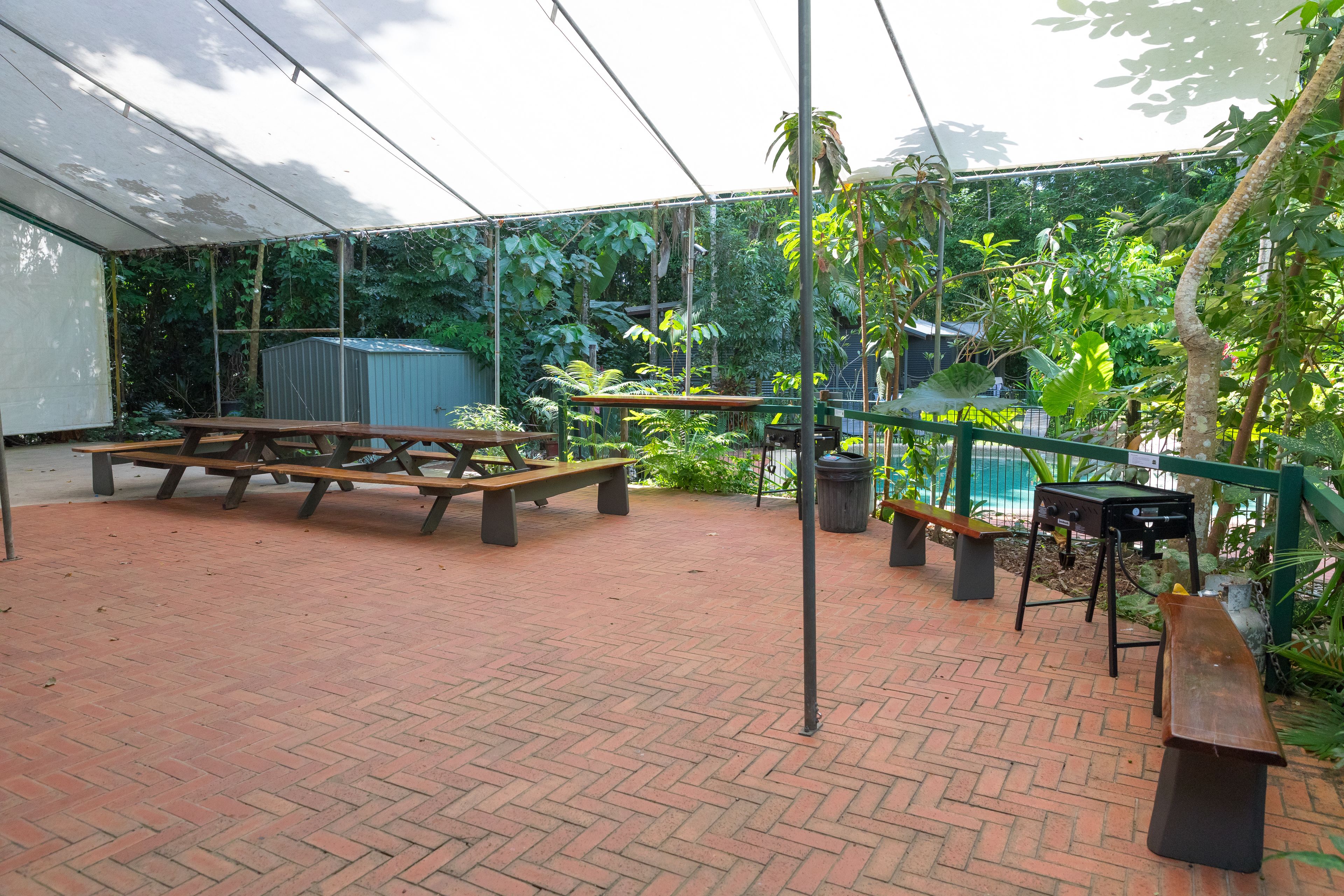 A covered patio area with picnic tables, a barbecue grill, and a pool in the background.