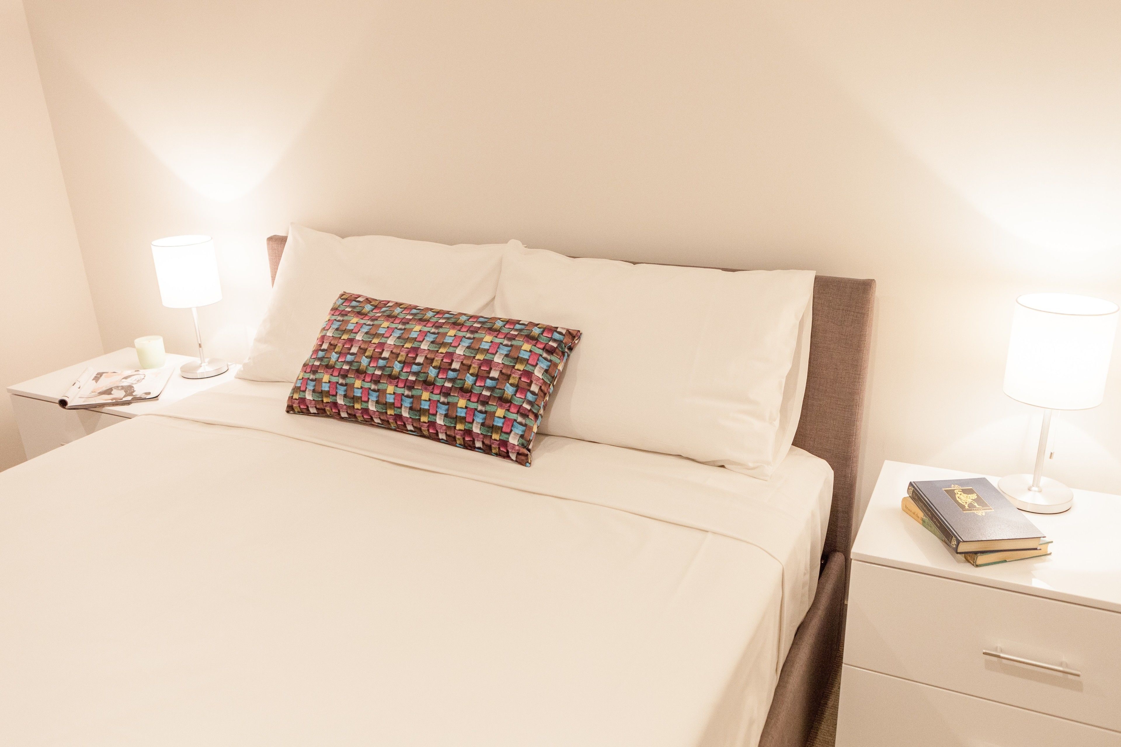 Een net opgemaakt bed met een kleurrijk kussen, een nachtkastje met een boek en een lamp.
