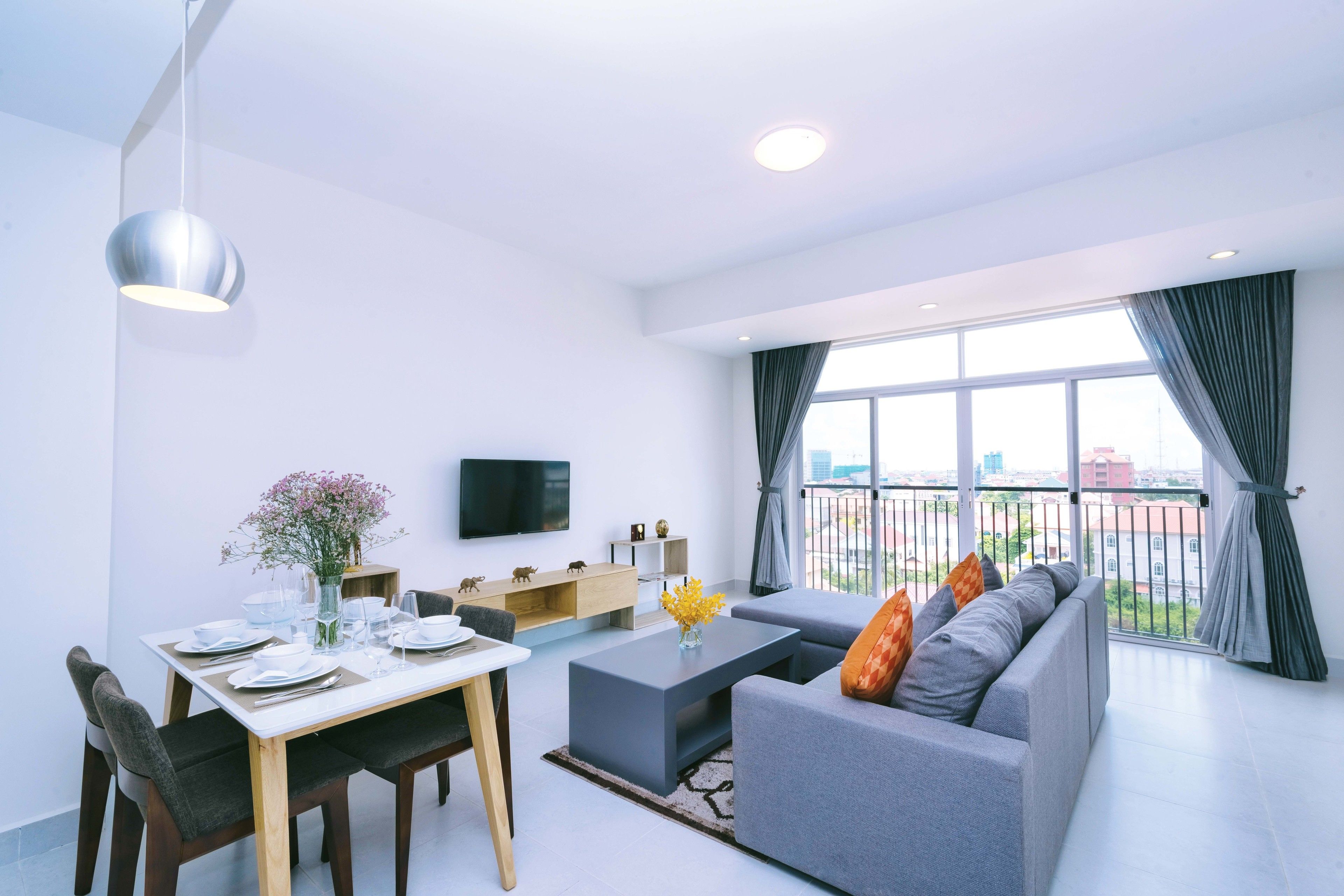 A modern living room with a dining area, a grey sofa, a flat-screen TV, and a large window with curtains.