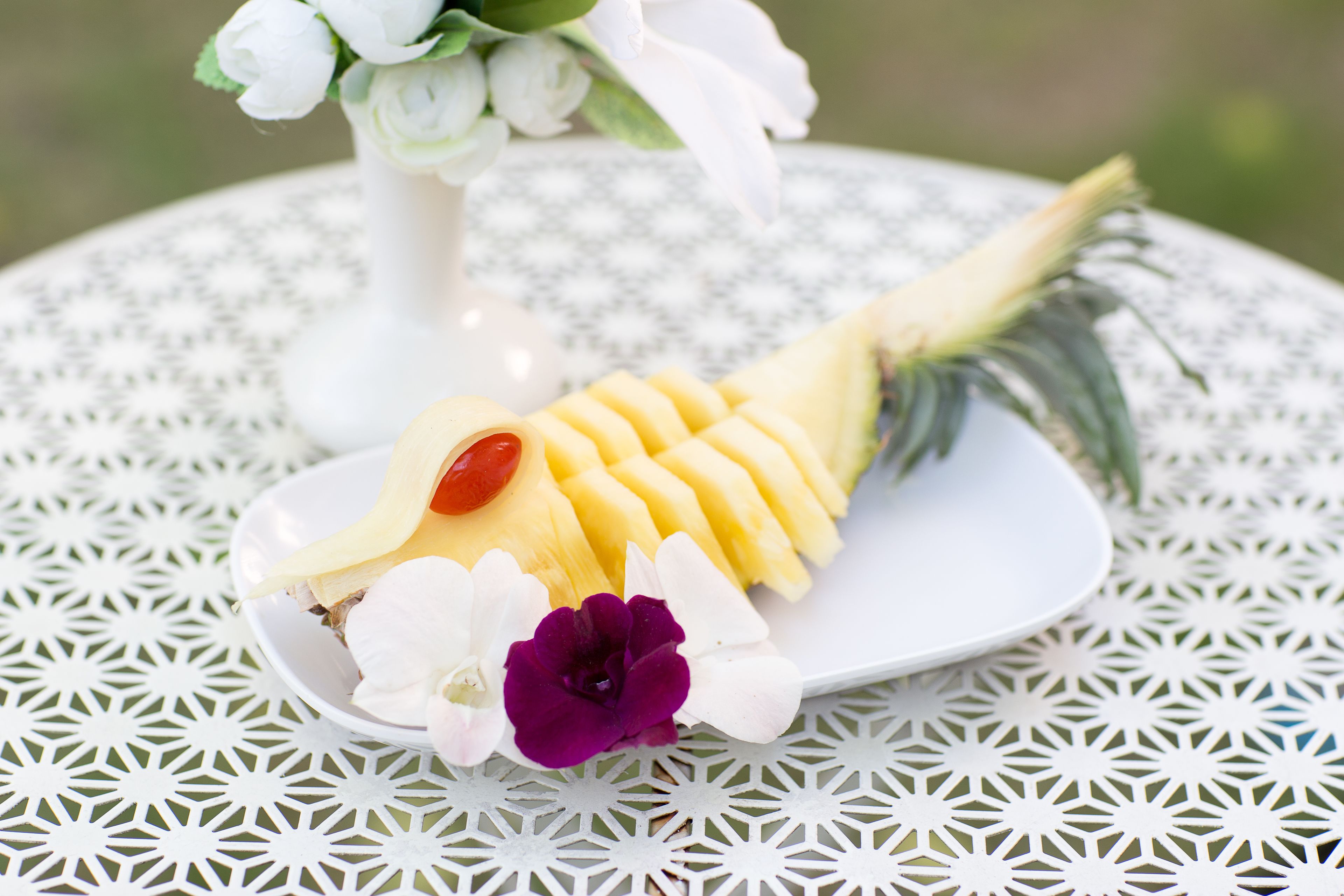 A white plate with a pineapple slice, a red cherry, and white and purple flowers on a white table with a geometric pattern.