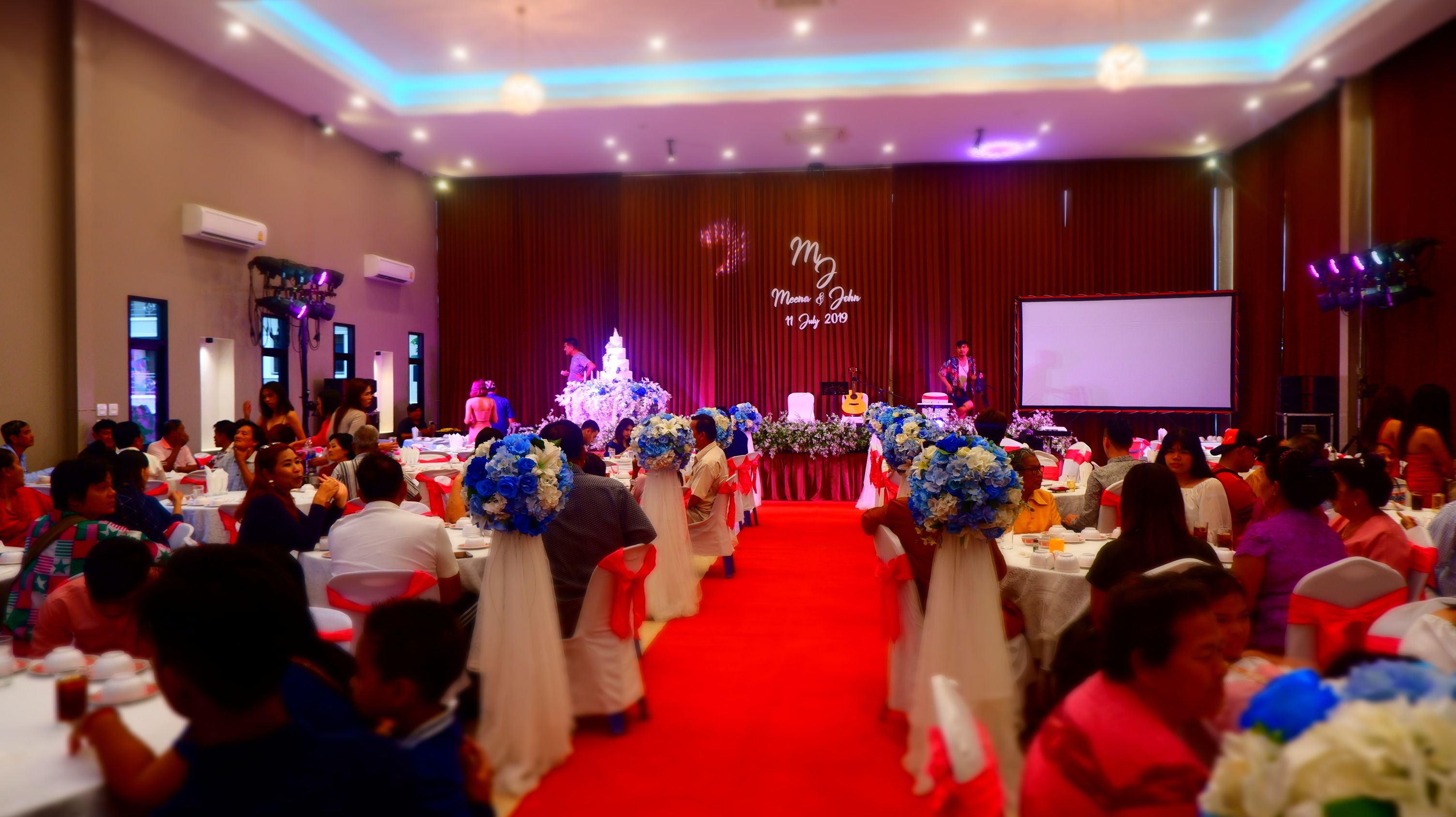 A banquet hall with round tables, a stage with floral decorations, and a red carpet.