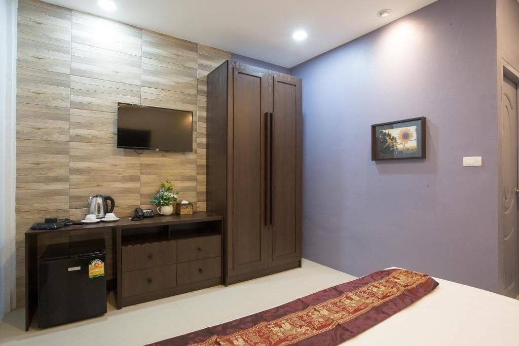 A bedroom with a bed, a wardrobe, a television mounted on the wall, a small desk with a kettle, a refrigerator, and a framed picture on the wall.