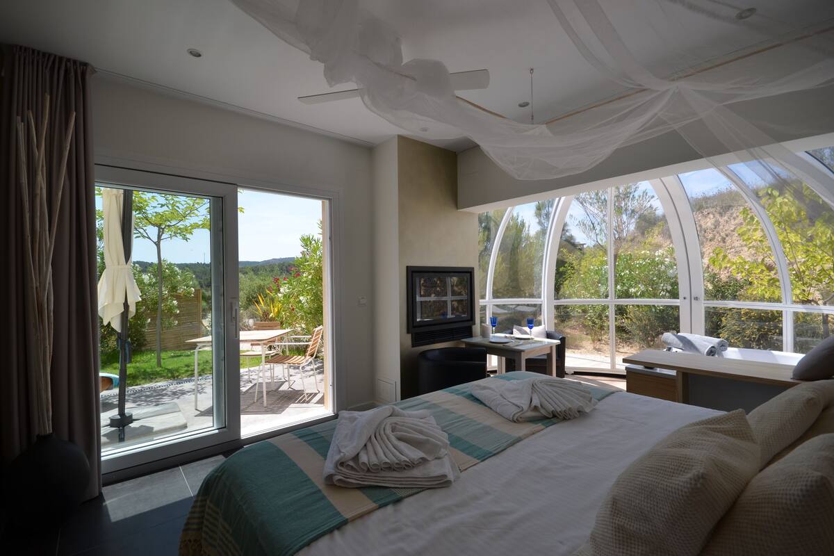 A modern hotel room with a large bed, a flat-screen TV, and a balcony with a view of the outdoors.