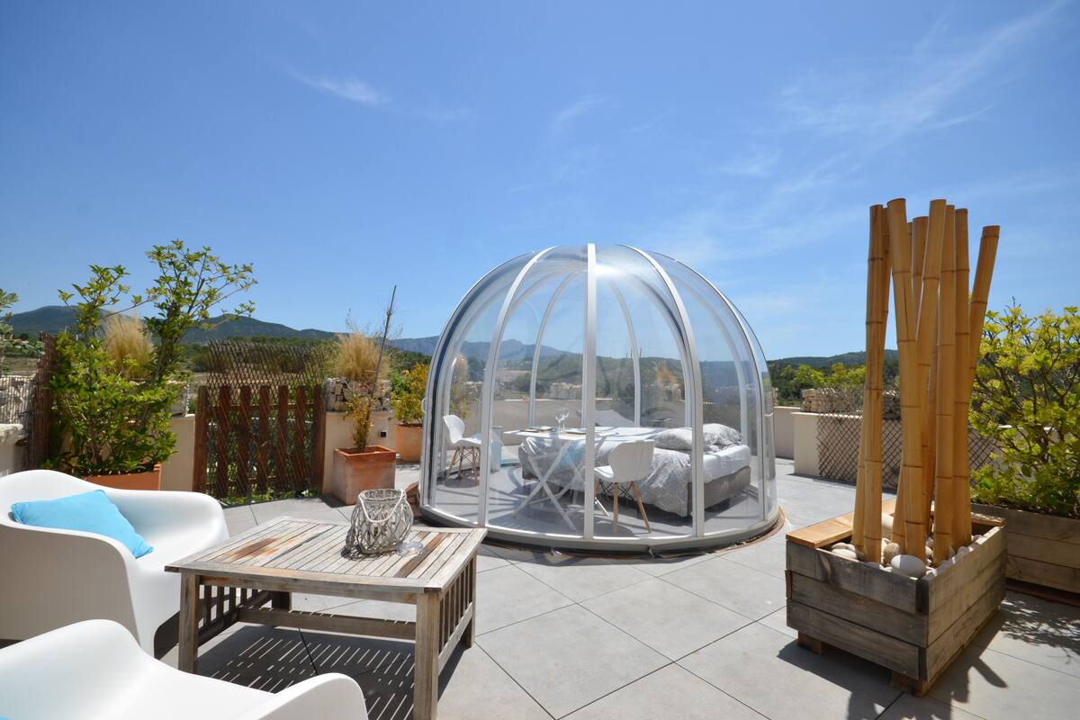 A transparent dome structure with a bed inside, surrounded by outdoor furniture and plants on a patio.