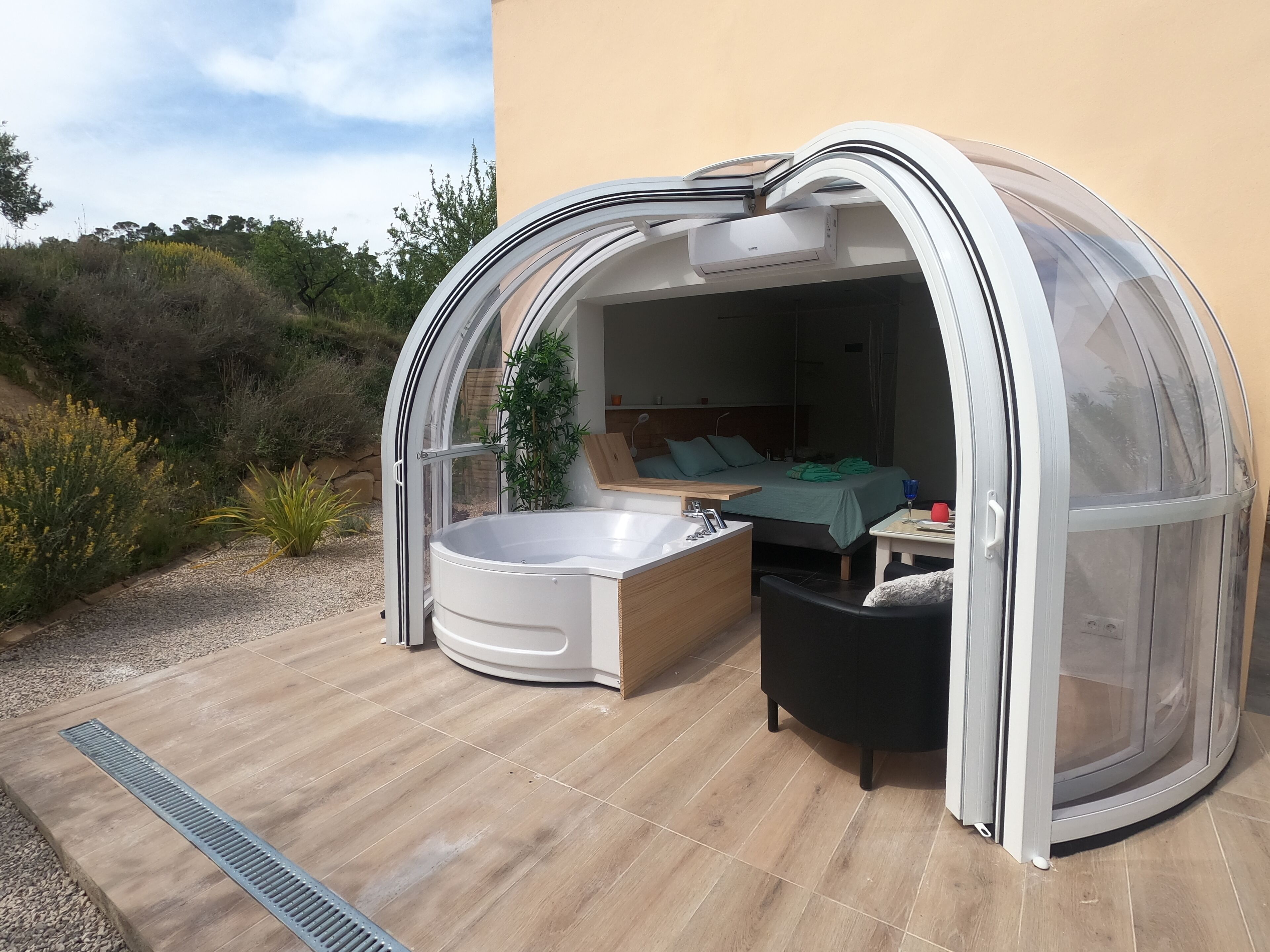 A modern outdoor bedroom with a glass enclosure, a hot tub, and a wooden bench.