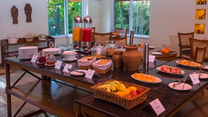 A buffet setup with various food items and drinks, including fruits, vegetables, and beverages, arranged on a glass table with chairs around it. - Dvara Siruvani (Coimbatore)