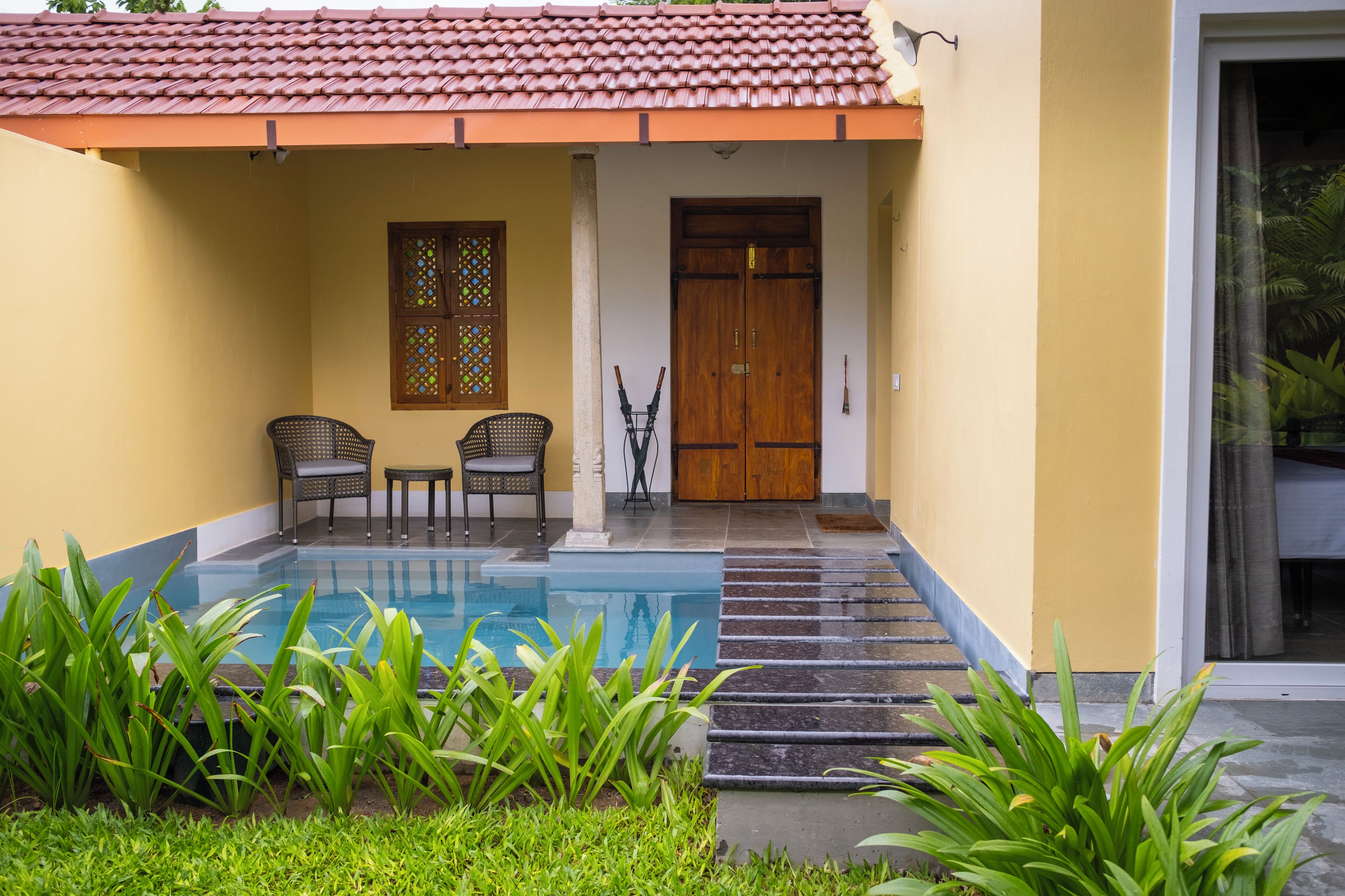 A pool with a stone-lined edge, two wicker chairs, a wooden door, and a window with a decorative pattern.