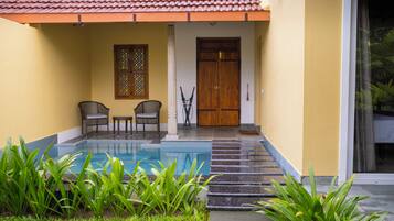 A pool with a stone-lined edge, two wicker chairs, a wooden door, and a window with a decorative pattern.