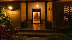 A hotel entrance with a sign reading "Kuvilam," a well-lit pathway leading to the interior, and a traditional architectural style with columns and a wooden door. - Dvara Siruvani (Coimbatore)