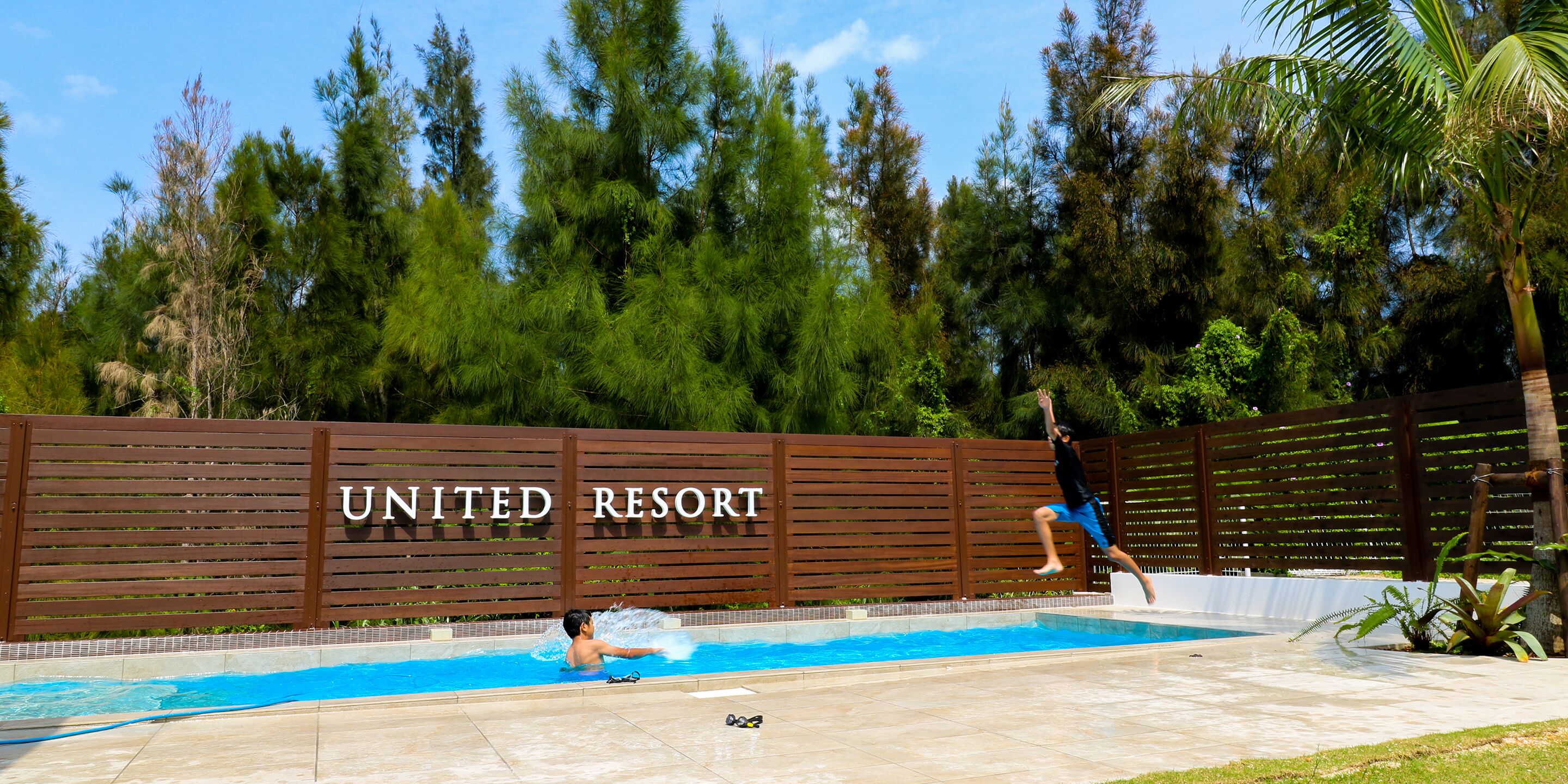 A swimming pool area with a wooden fence labeled "UNITED RESORT" and a palm tree.