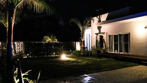 Een nachtzicht van een huis met een verlichte voortuin, een palmboom en een rieten dakconstructie.