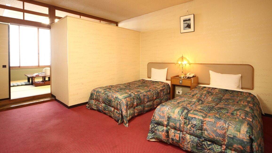 A hotel room with two beds, a red carpet, a wooden headboard, a nightstand with a lamp, and a window with a view.