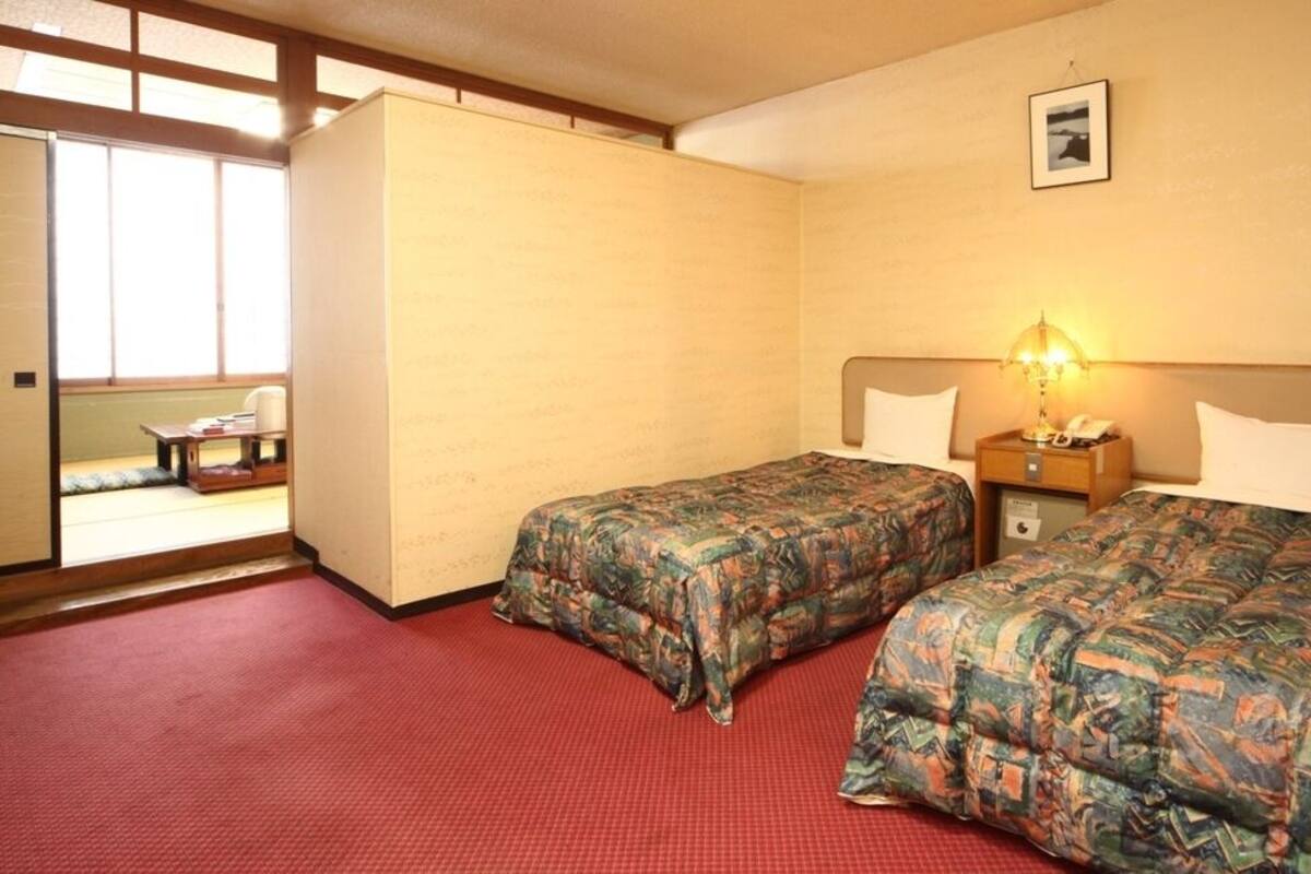 A hotel room with two beds, a red carpet, a wooden headboard, a nightstand with a lamp, and a window with a view.