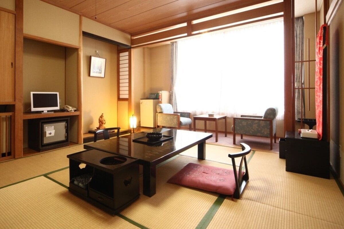 Una habitación japonesa tradicional con suelo de tatami, una mesa baja, un televisor, un refrigerador y una puerta corredera que da a un balcón.