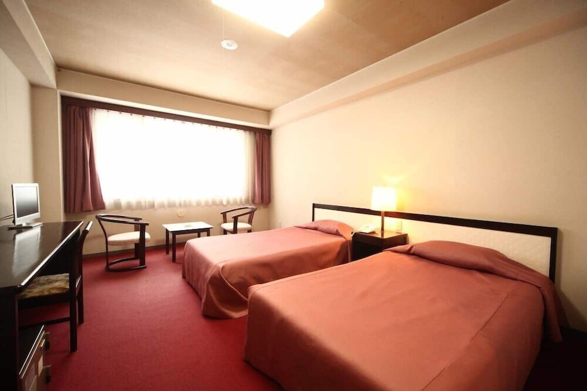 A hotel room with two beds, a desk, a chair, a lamp, and a window with curtains.