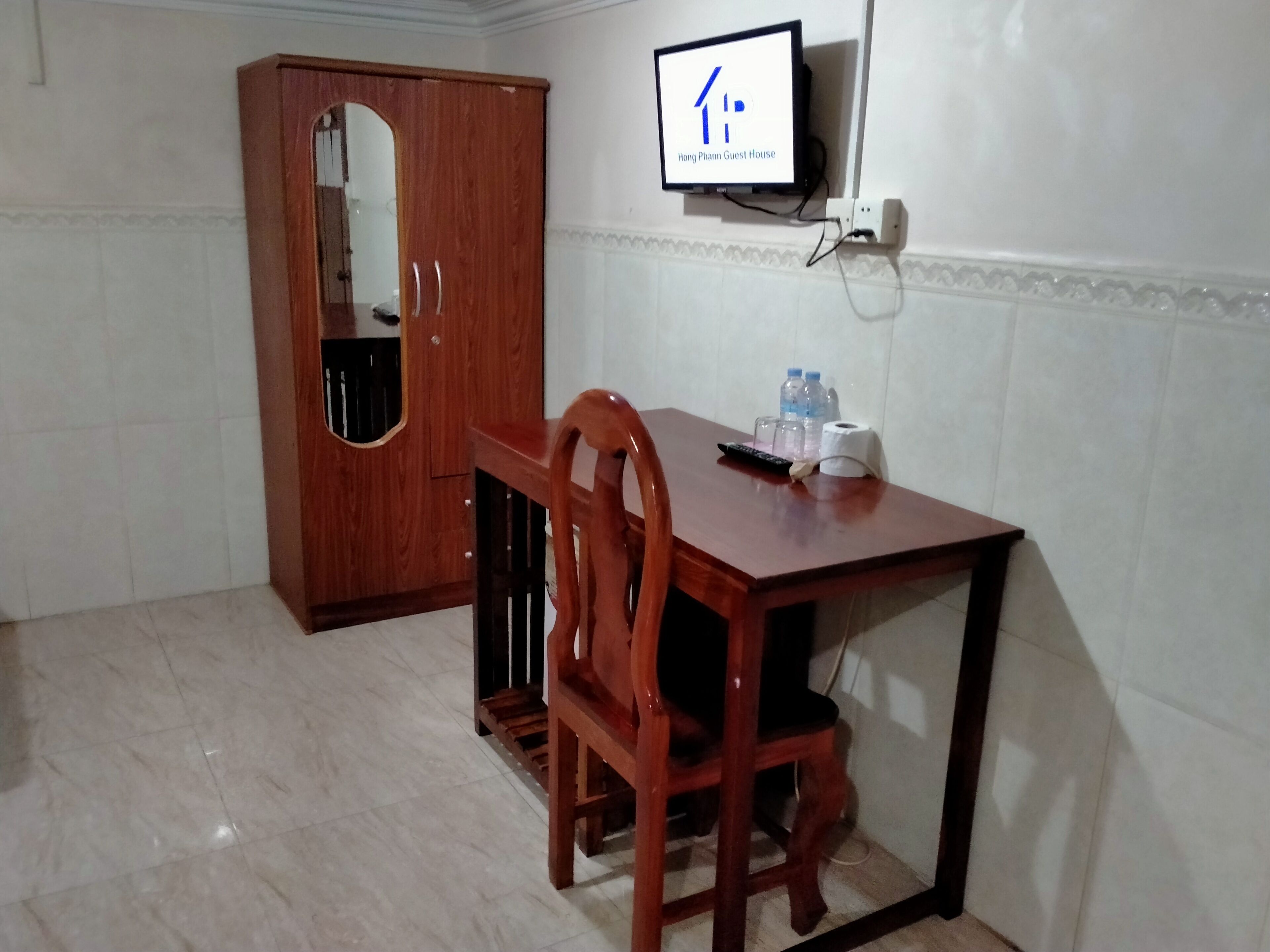 Een kamer met een houten kast, een houten eettafel met stoelen, een aan de muur bevestigde televisie en een kleine tafel met water in flessen en een afstandsbediening.