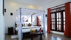 A four-poster bed with a canopy, a bedside table with a fruit basket, and a window with red curtains. - Hotel Sanmark (Ahangama)