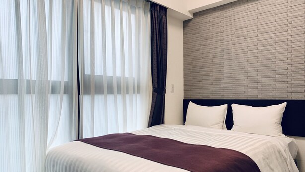 Quarto de hotel moderno com uma cama grande, cabeceira em padrão de tijolos e cortinas leves.